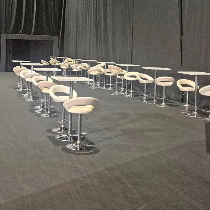 Image of A set of modern bar stools with curved cream cushions and chrome bases, paired with matching square high-top tables, arranged on a dark stage in front of a black curtain.