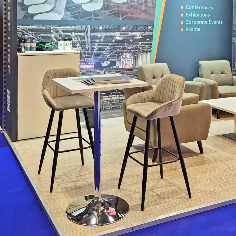Image of A modern booth display featuring a square high-top table with a chrome pedestal base and two upholstered bar stools, alongside matching lounge armchairs in a conference-exhibition setting.