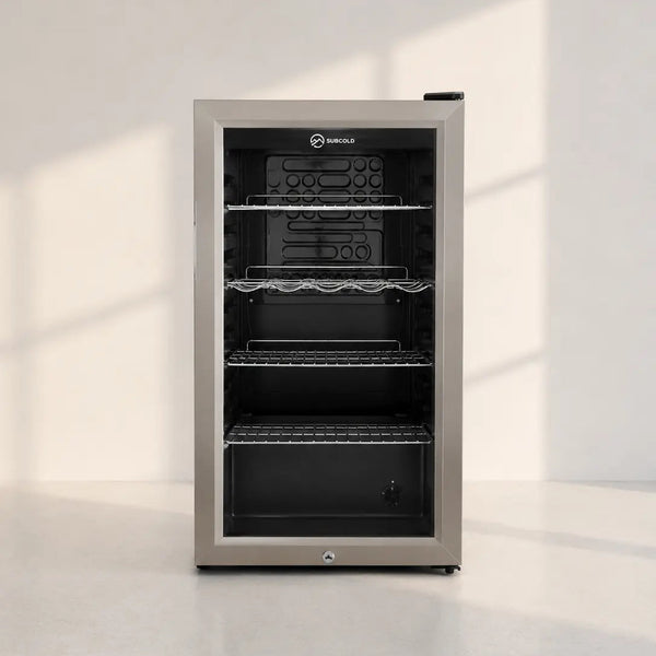 A single-door undercounter beverage refrigerator with a brushed stainless steel exterior and a black glass door, shown on a light gray floor against a white wall with soft window shadows.