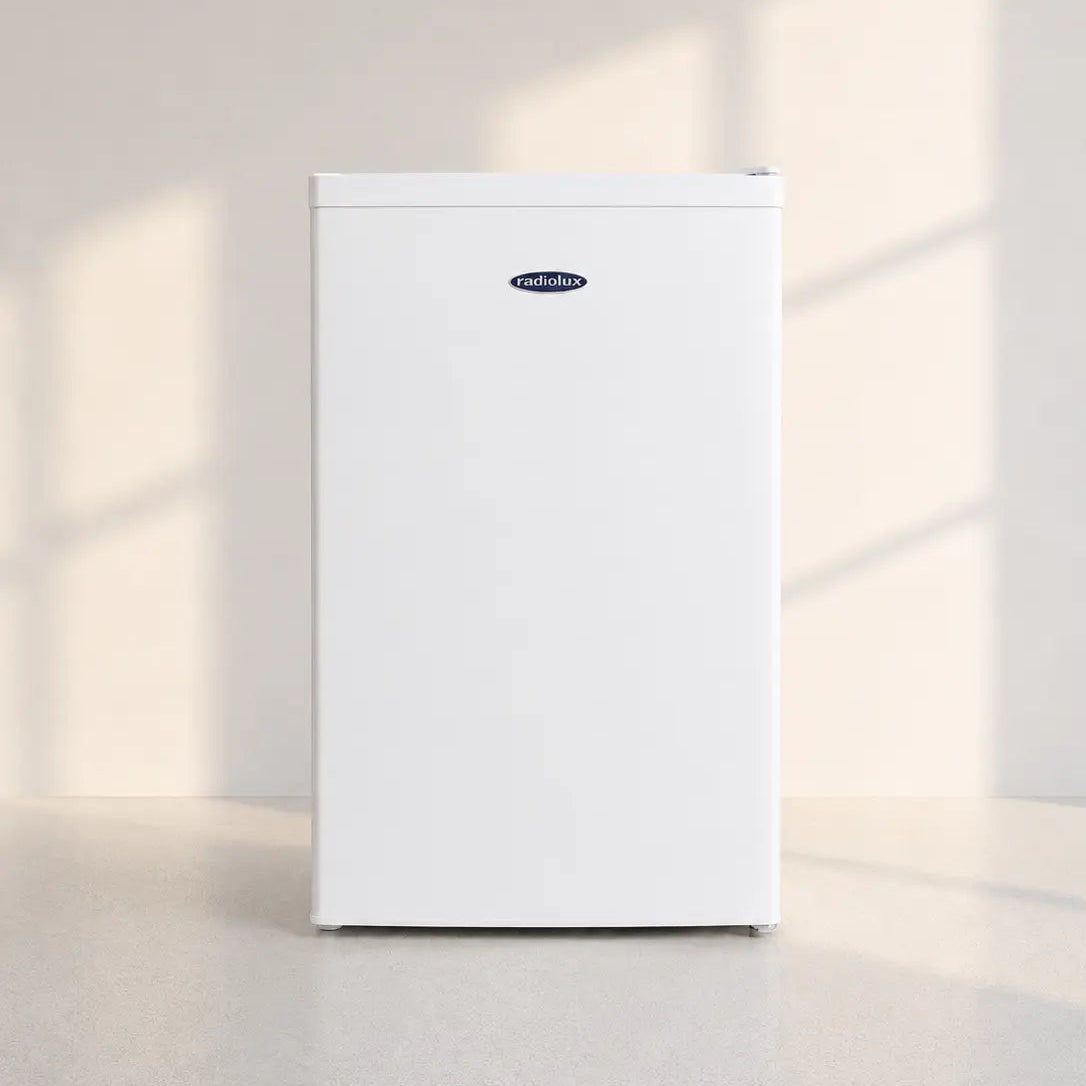 A white mini refrigerator with a small blue and white Frigidaire logo on the upper front, standing on a light gray floor against a beige wall with soft window shadow projections.