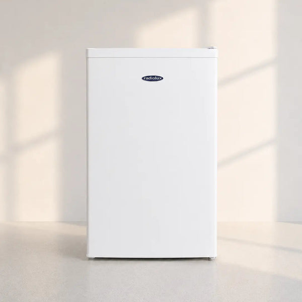 A white mini refrigerator with a small blue and white Frigidaire logo on the upper front, standing on a light gray floor against a beige wall with soft window shadow projections.