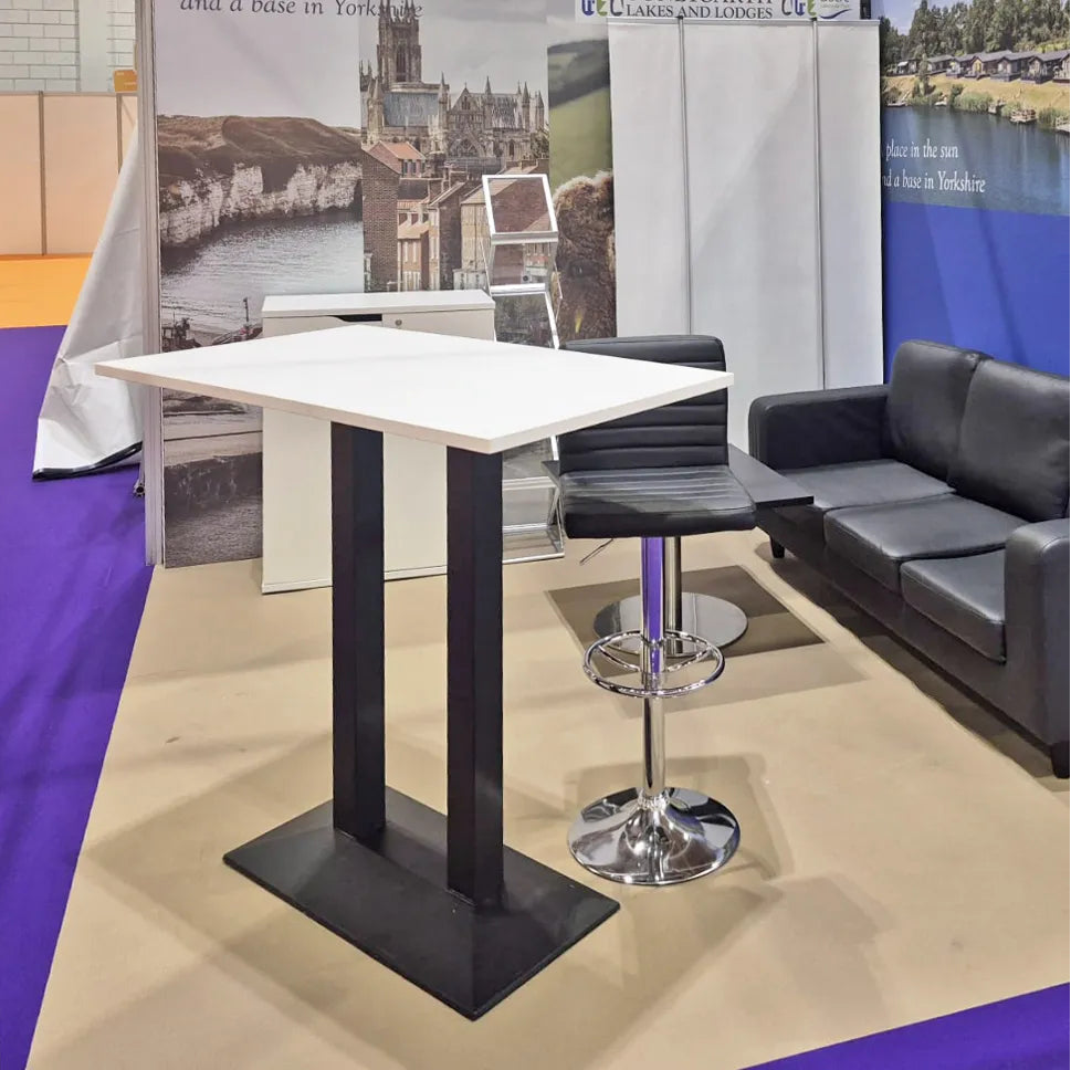 Image of A modern high-top bar table with a white rectangular top and black metal pedestal legs, shown in a trade show booth next to a black leather sofa and a chrome swivel barstool.