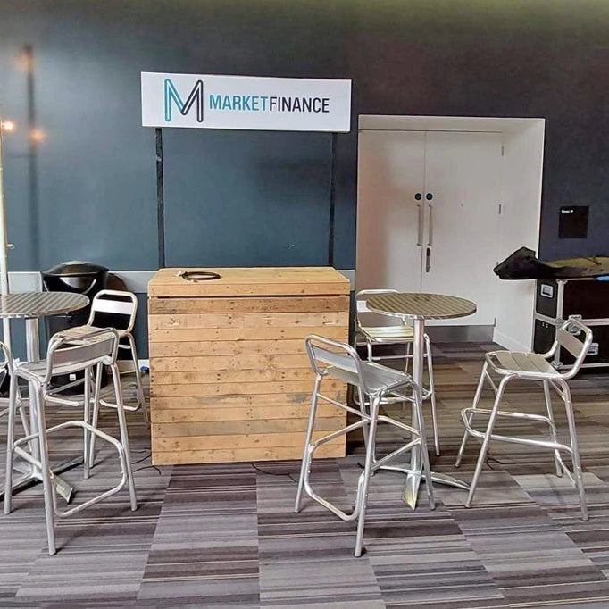 Image of A styled event or lounge setup featuring a central wooden bar counter, four silver metal bar stools, two small round tables, and a large black equipment case against a dark blue-gray wall with a 'MARKETFINANCE' sign.