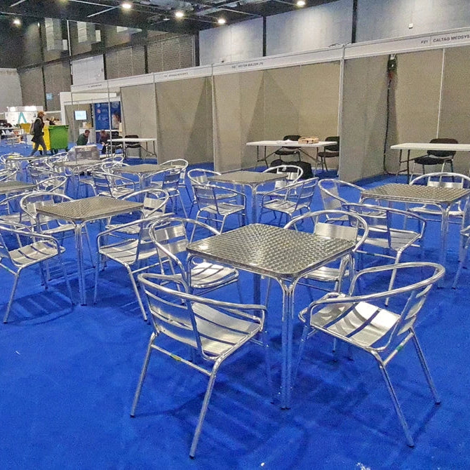 A large exhibition hall floor covered in bright blue carpet, with many small metal tables and chairs arranged in rows. White booth walls and other booths are visible in the background, along with a few people and objects in the distance.