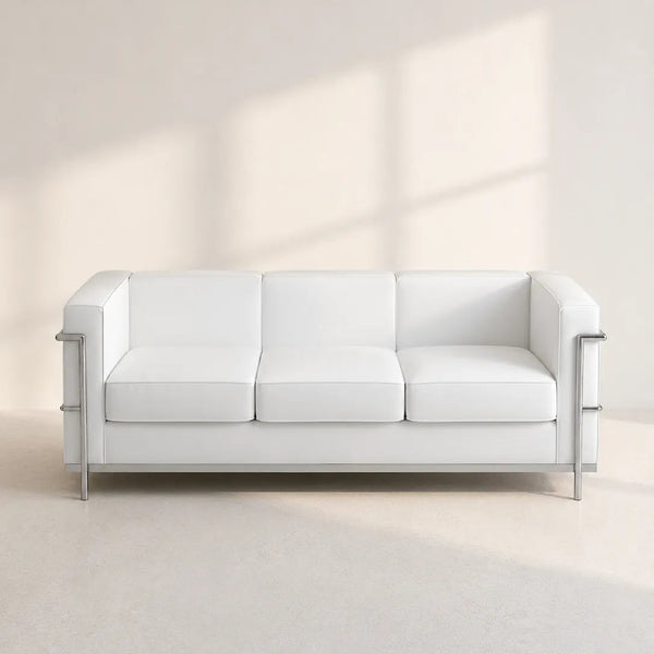 A modern white leather three-seat sofa with slim silver metal legs and side accents, set against a minimalist beige wall with soft window shadow projections.