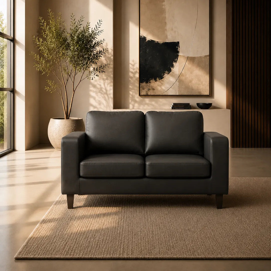 Image of A modern two-seat sofa in dark charcoal gray leather, positioned in a minimalist, warm-toned living room with a large potted plant, abstract wall art, and a textured rug.