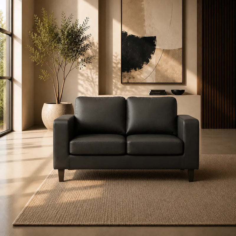 A modern two-seat sofa in dark charcoal gray leather, positioned in a minimalist, warm-toned living room with a large potted plant, abstract wall art, and a textured rug.