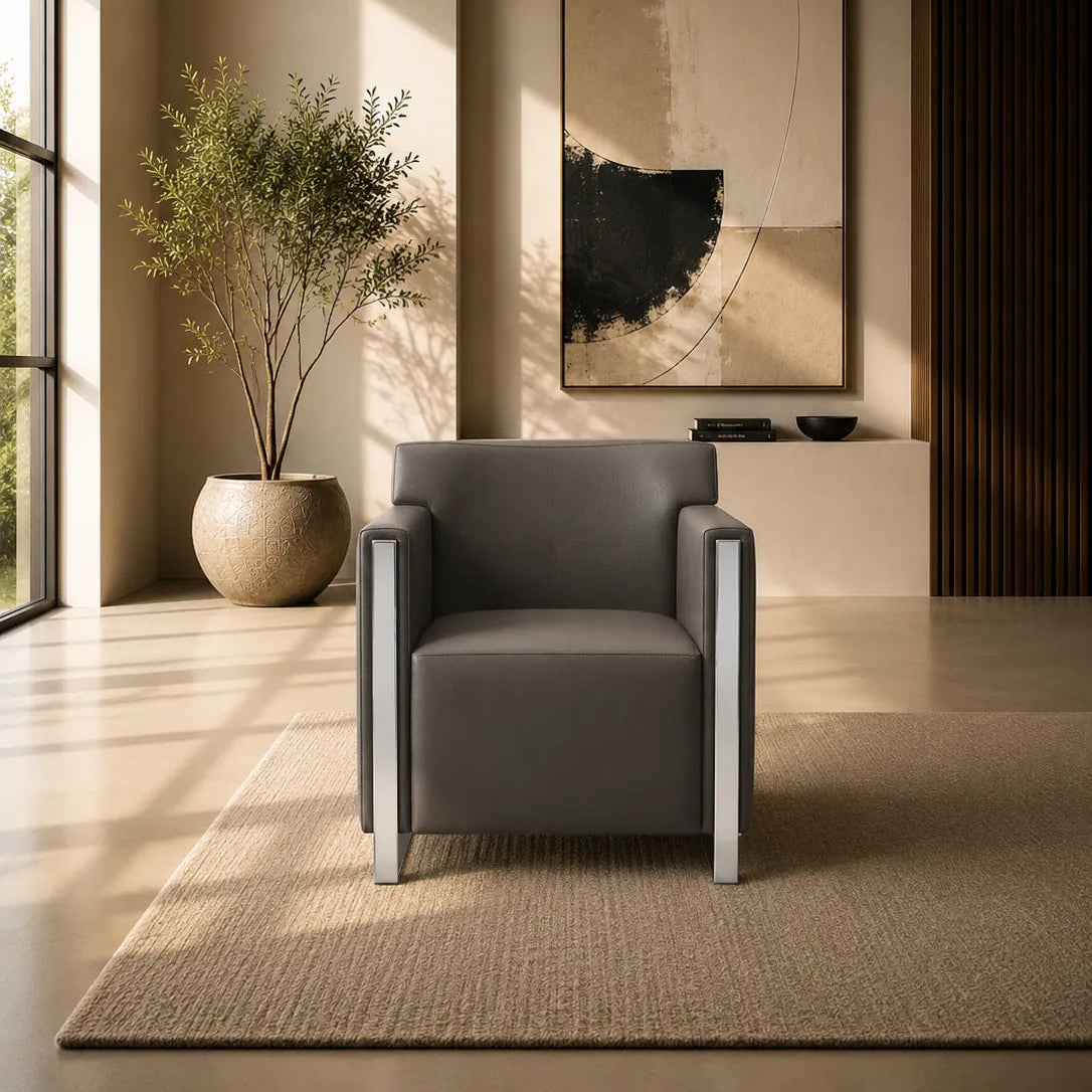 A modern gray upholstered armchair with slim silver metal legs sits on a textured beige rug in a sunlit minimalist room. A large abstract wall art piece hangs above a small console, with a potted plant near a window and warm ambient lighting.