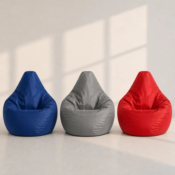 Three teardrop-shaped bean bag chairs in blue, gray, and red, lined up side-by-side on a light gray floor against a beige wall with soft window shadows.
