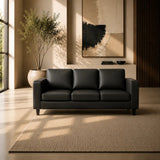 A modern three-seater sofa in black leather sits in a minimalist, sunlit living room with beige walls, a large abstract painting, a potted plant, and a textured beige rug.