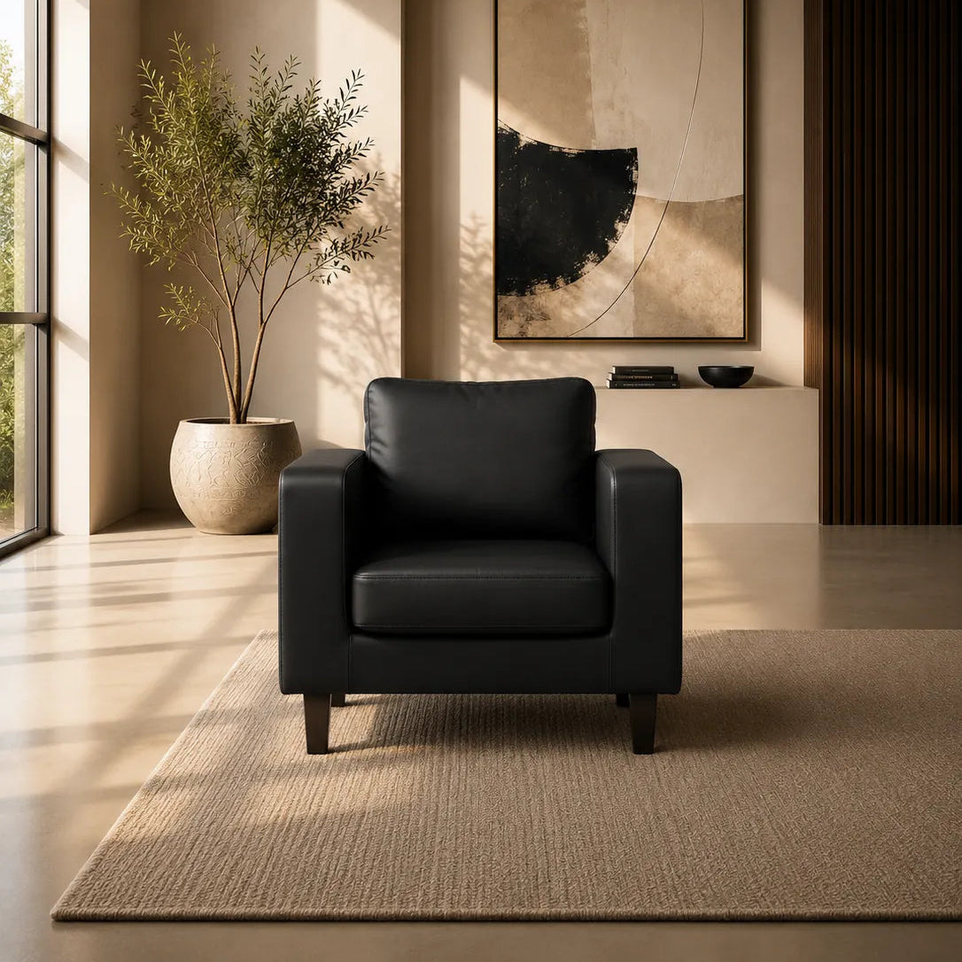 A modern black leather armchair in a minimalist, warm-toned living room setting with a large potted plant, abstract wall art, and a textured rug.
