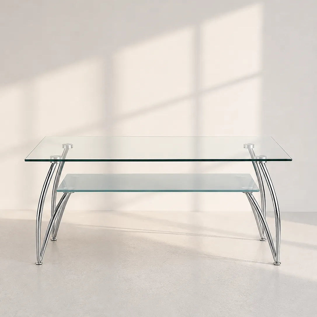 A modern rectangular glass coffee table with a clear glass top and lower shelf, supported by curved silver metal legs, set against a minimalist off-white wall with soft window shadow projections.