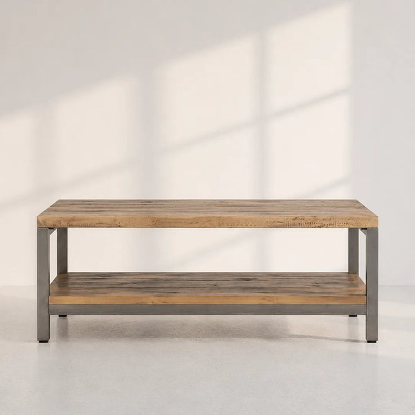 A rectangular coffee table with a rustic light brown wood top and lower shelf, supported by a gray metal frame, set against a light gray wall with soft window shadow patterns.