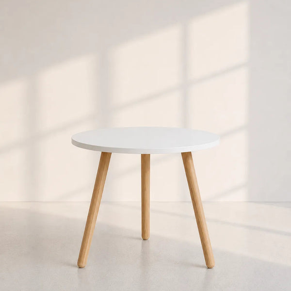 A minimalist round side table with a white top and light natural wood legs, set against a warm beige wall with soft window shadow patterns.