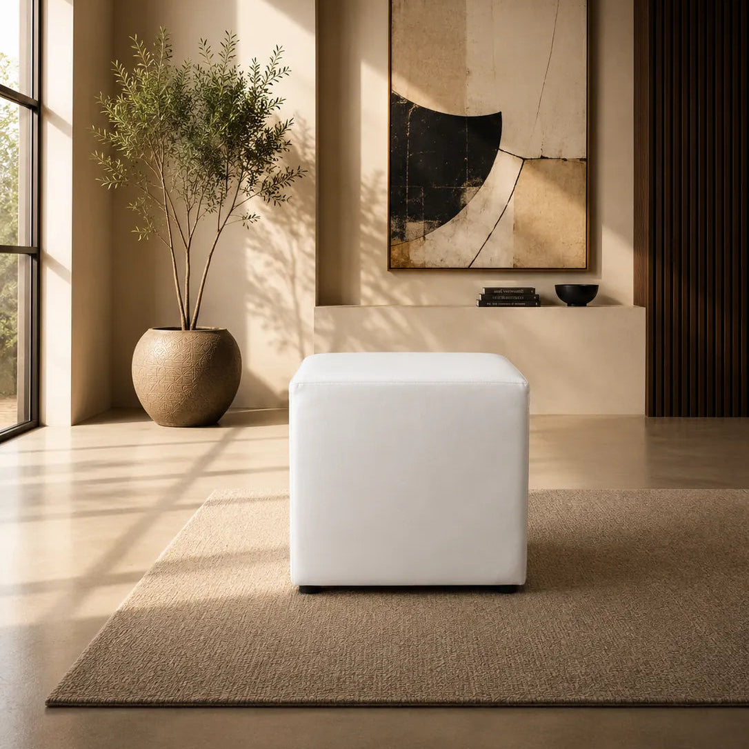 Image of A minimalist modern interior featuring a white cube ottoman on a textured beige rug, with a large abstract art print on the wall behind it, a potted plant, and warm sunlight streaming through a window.