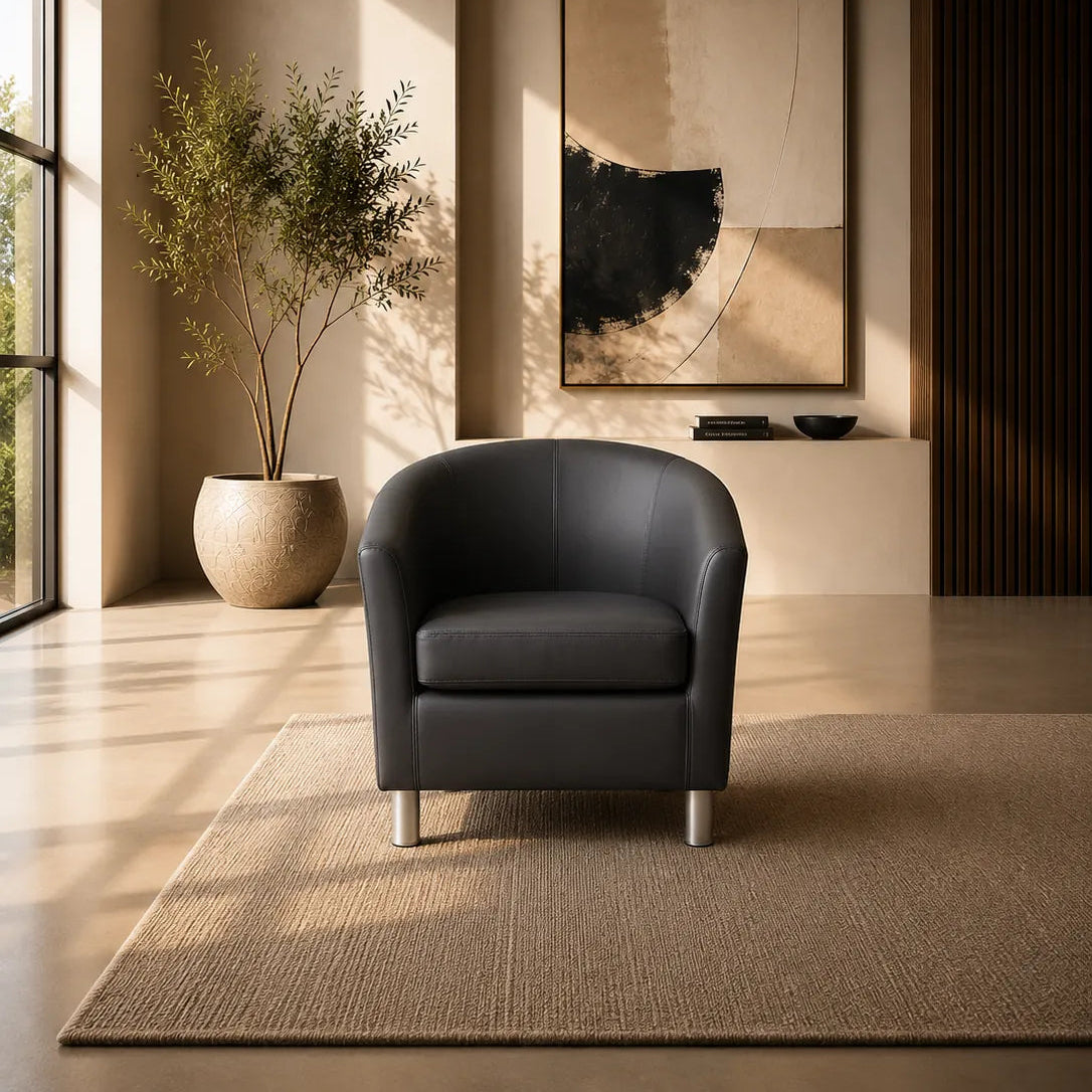 A modern dark gray leather armchair with silver legs sits on a textured beige rug in a sunlit minimalist living room. A large abstract black, beige, and white framed print hangs on the wall behind it, with a potted plant to the left.