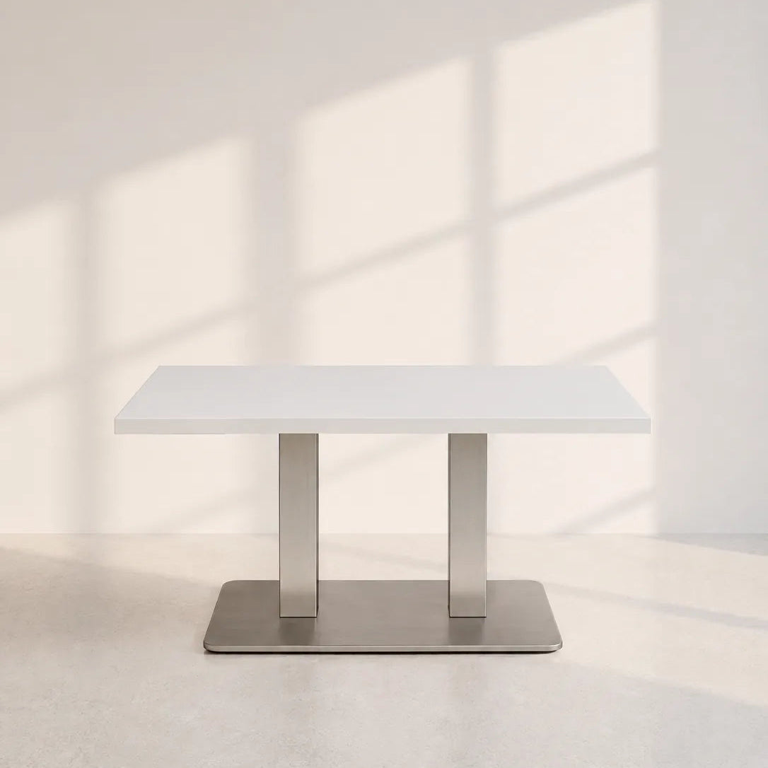 Image of A modern rectangular dining table with a white top and a brushed silver metal pedestal base, set against a minimalist off-white wall with soft window shadow patterns.