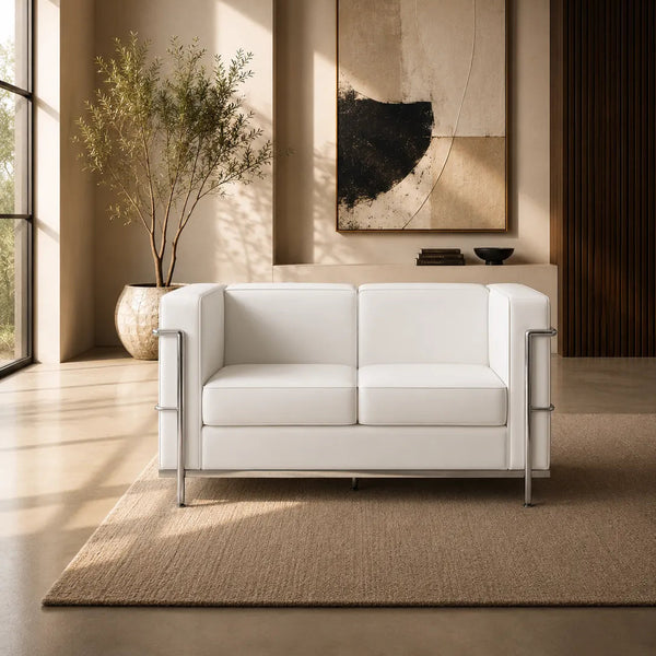 A modern white two-seat sofa with slim silver metal legs, positioned on a textured beige rug in a minimalist, warm-toned living room with a large abstract art print on the wall.