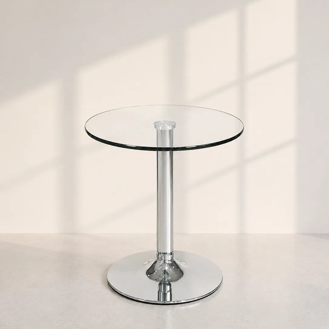 Image of A modern round glass-top pedestal table with a polished chrome base, set against a warm off-white wall with soft window shadow patterns.