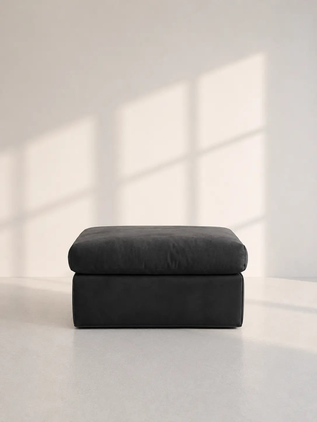 Image of A dark charcoal gray upholstered ottoman with a square cushioned top and a thick rectangular base, set against a minimalist off-white wall with soft window shadow patterns.