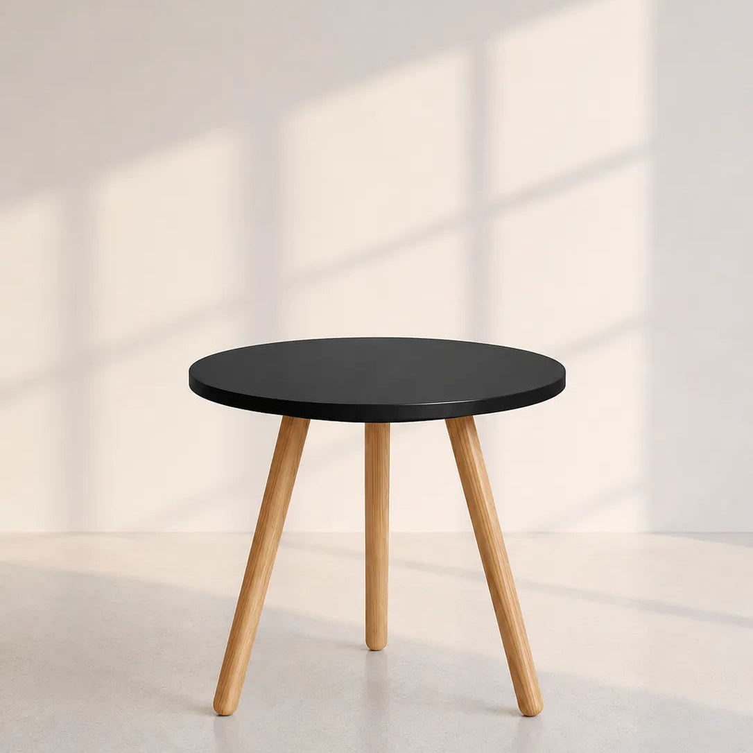 Image of A modern round side table with a matte black top and three tapered natural wood legs, set against a minimalist off-white wall with soft window shadow patterns.