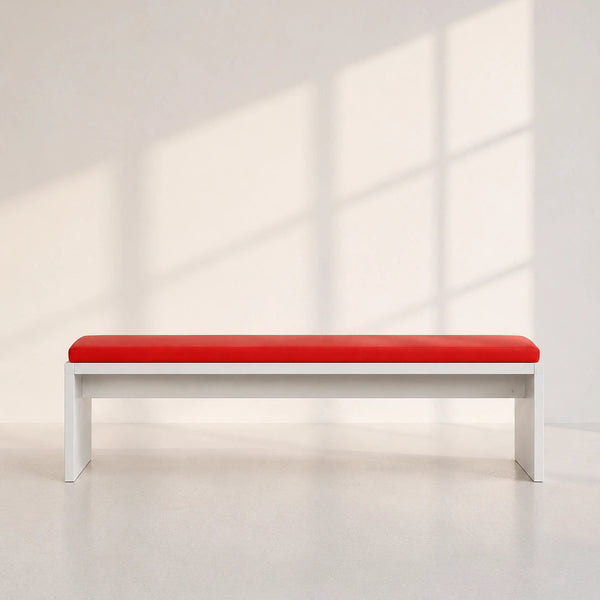 A minimalist bench with a bright red upholstered seat and a white frame, positioned against a light gray wall with soft window shadow patterns.