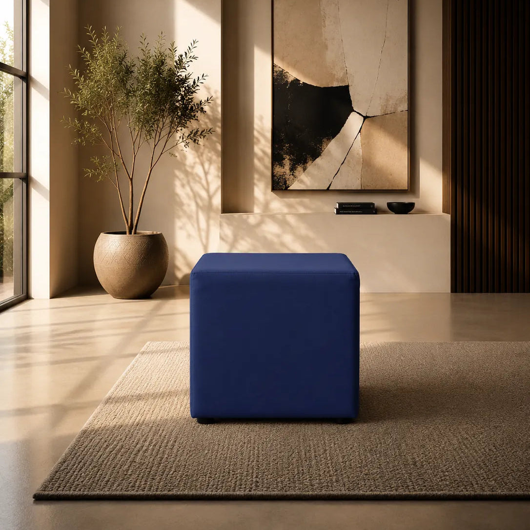 Image of A modern blue cube ottoman or bench placed on a textured beige rug in a sunlit minimalist interior with a large potted plant, wall art, and warm ambient lighting.