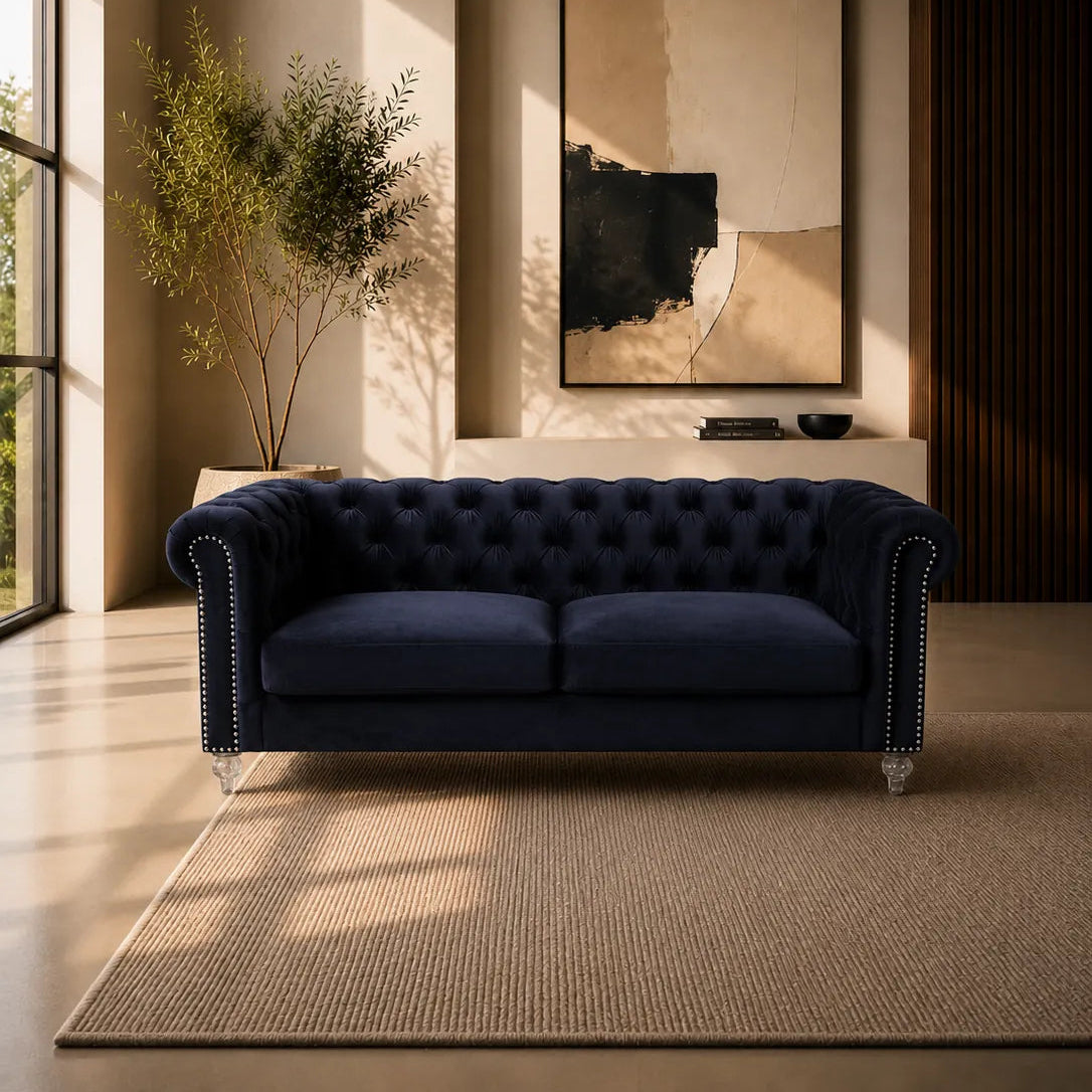 A navy blue tufted velvet sofa with silver nailhead trim and clear glass legs, placed in a modern living room setting on a beige rug with a large abstract painting on the wall behind it.