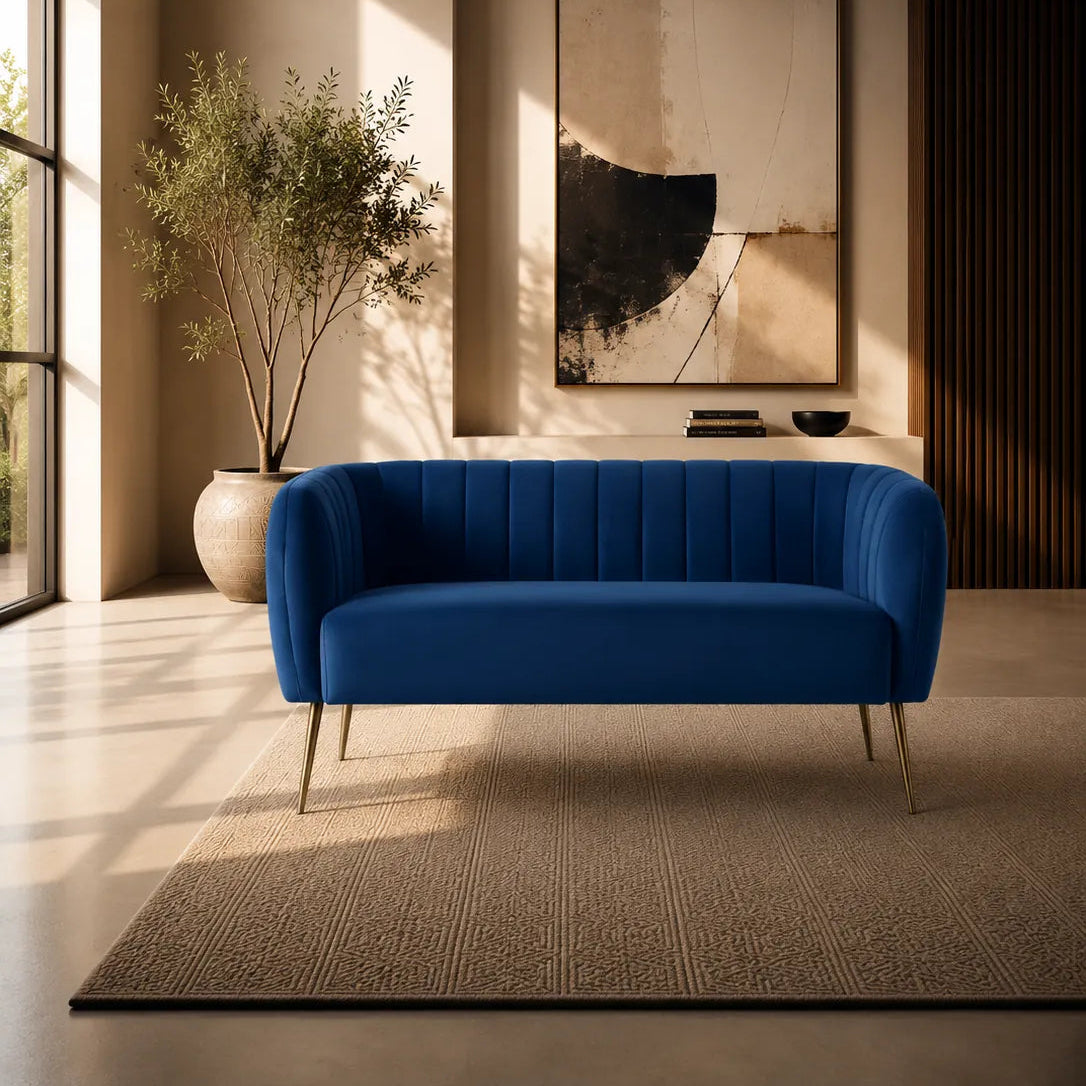 Image of A modern blue tufted loveseat with slim gold legs, positioned on a textured beige rug in a contemporary living room setting with warm lighting and an abstract wall art piece.