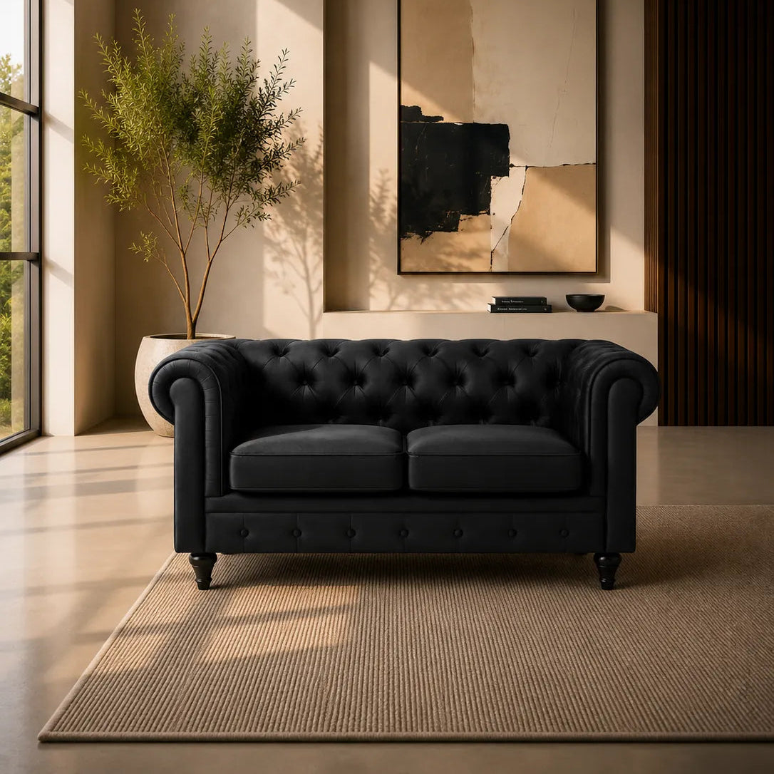 Image of A black tufted Chesterfield-style sofa placed in a modern living room with warm sunlight, a large abstract wall art piece, a potted plant, and a textured rug.