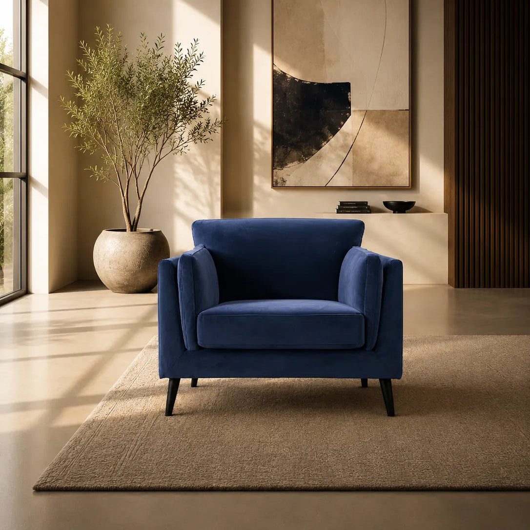 A modern blue velvet armchair with dark tapered legs, placed on a beige rug in a minimalist, warm-toned living room setting with a potted plant and abstract wall art.