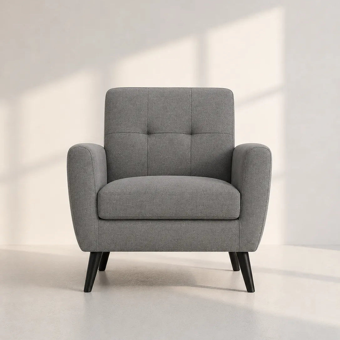 A modern gray upholstered armchair with a tufted backrest and tapered black legs, shown in a minimalist room with soft window light casting shadows on the wall.