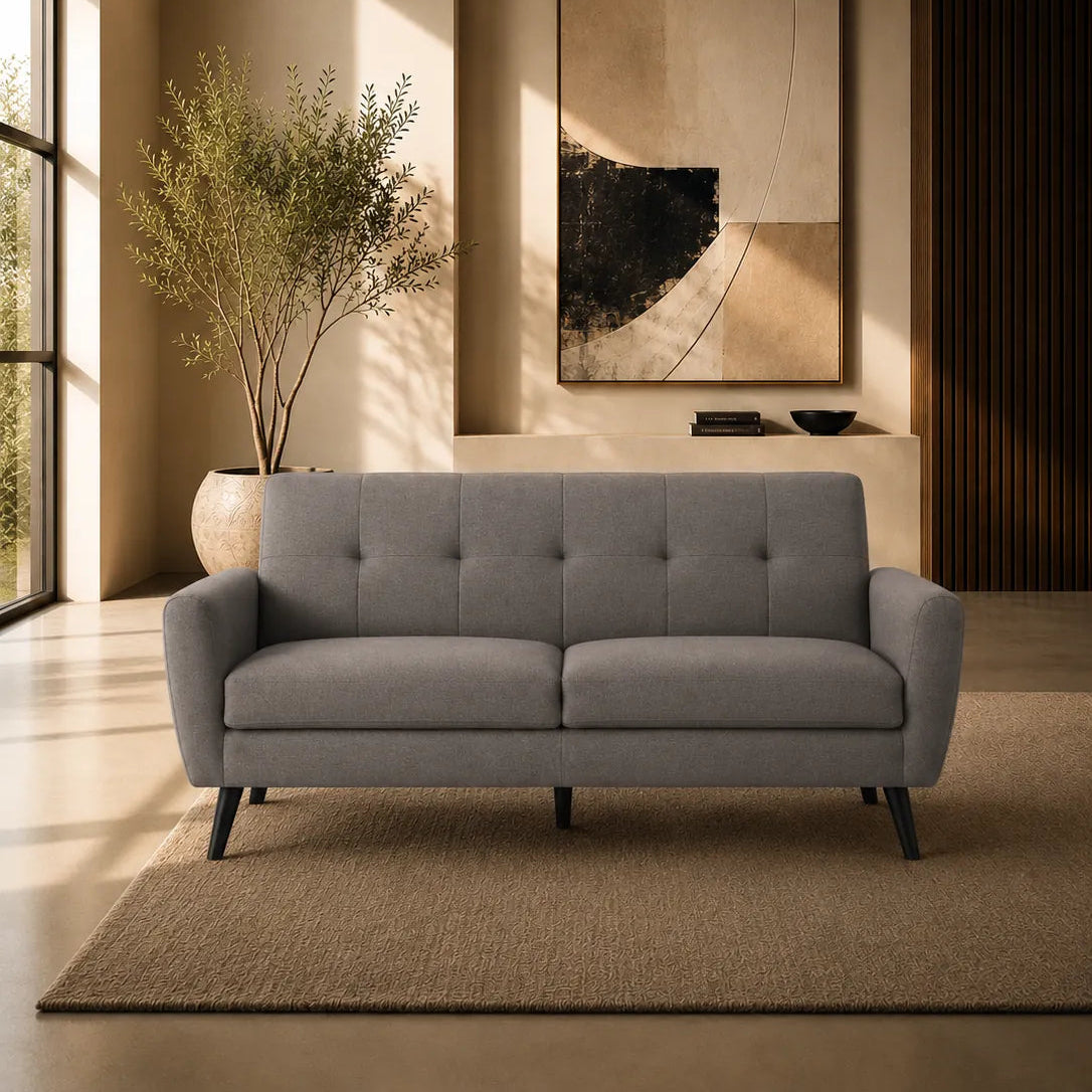 A modern light gray tufted loveseat sofa placed in a warm, minimalist living room with beige walls, a large abstract painting, a potted plant, and a textured rug.