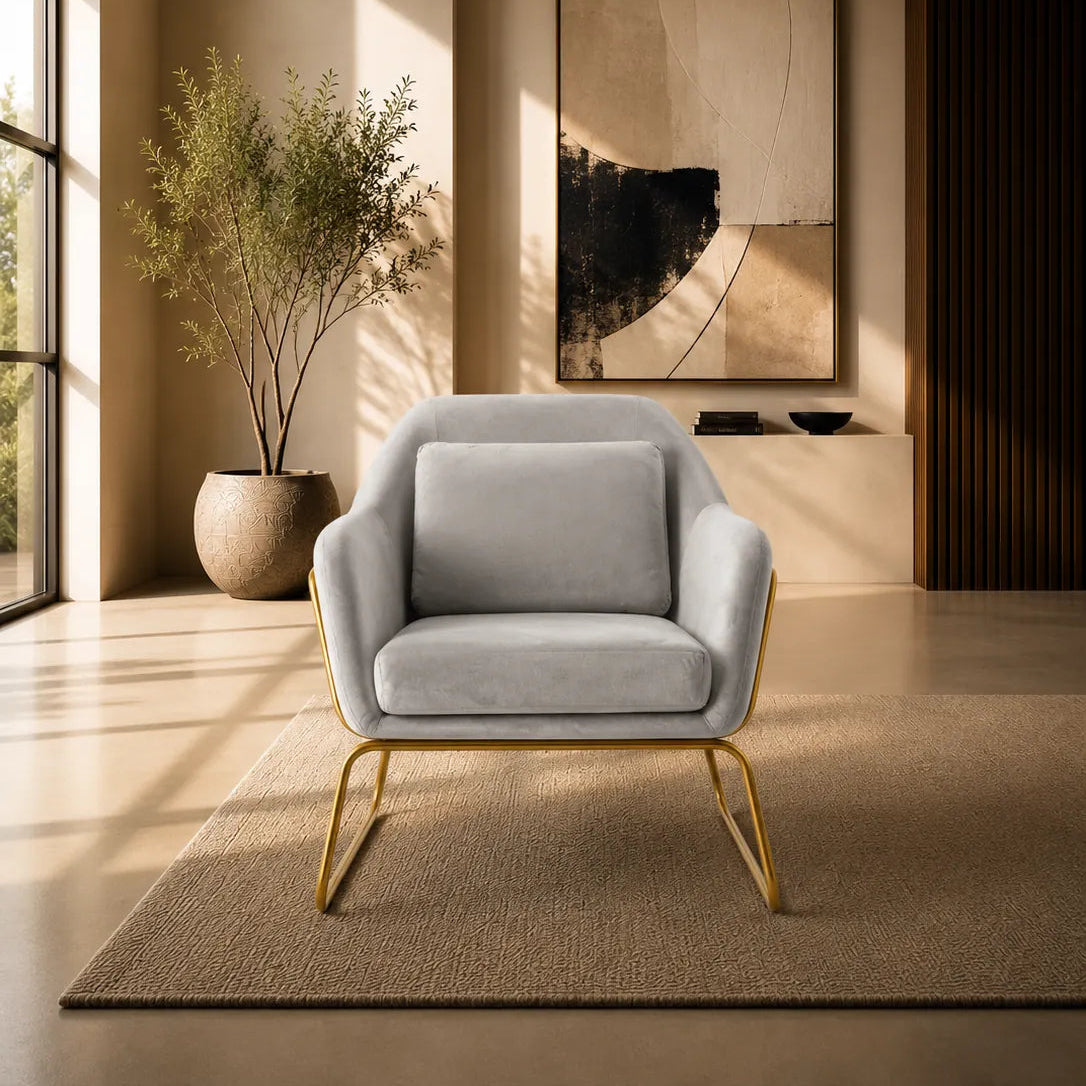 A modern light gray upholstered accent chair with gold metal legs, placed on a textured beige rug in a warm, sunlit contemporary living room.