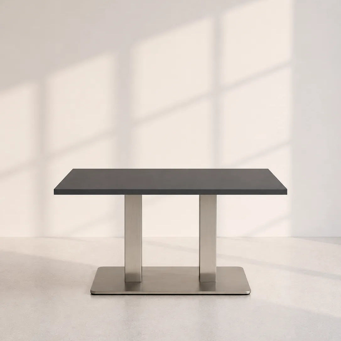 A modern rectangular dining table with a dark gray top and a brushed silver metal pedestal base, set against a minimalist off-white wall with soft window shadow patterns.