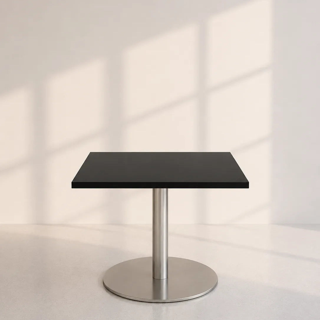 Image of A modern square table with a matte black top and a brushed silver metal pedestal base, set against a minimalist off-white wall with soft window shadow patterns.