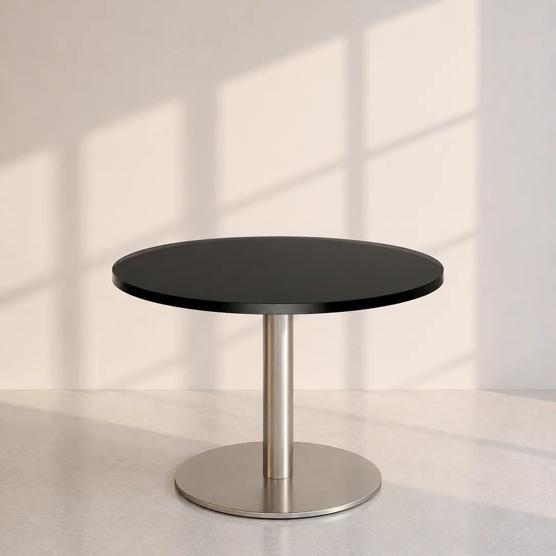 Image of A minimalist round table with a black top and a brushed silver metal pedestal base, set against a light gray wall with soft window shadow patterns.