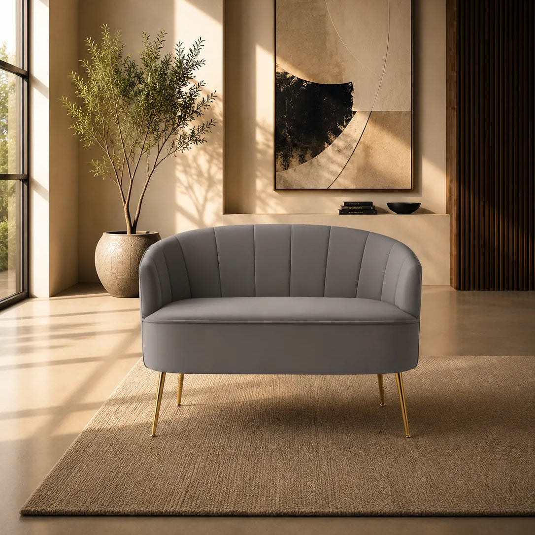 A modern gray upholstered lounge chair with gold legs, placed on a woven beige rug in a warm, sunlit interior room with a potted plant and abstract wall art.