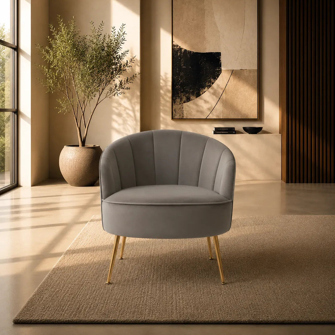 A modern gray upholstered accent chair with gold legs, placed on a textured beige rug in a warm, sunlit interior room with a potted plant and abstract wall art.