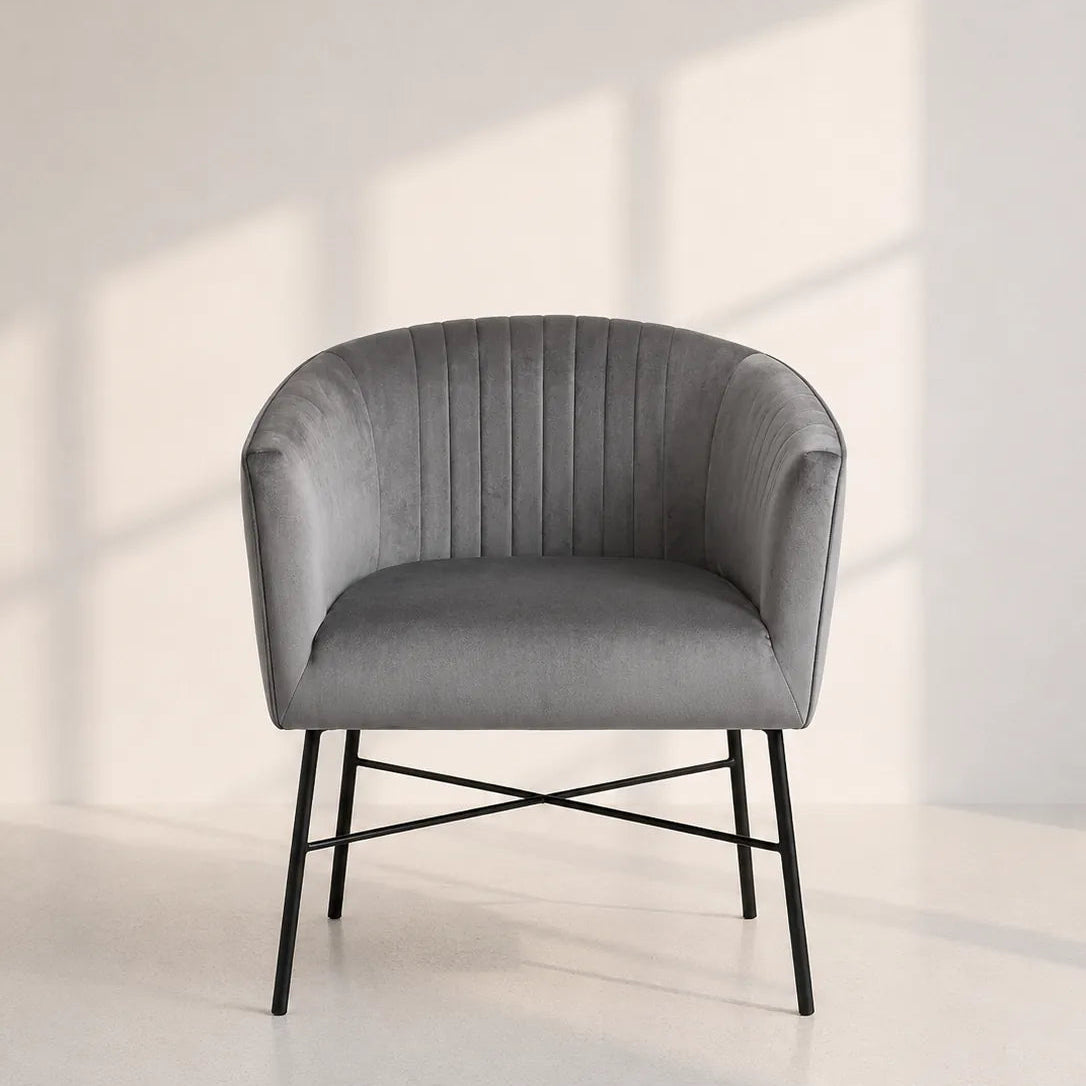 Image of A modern gray velvet armchair with a curved back, vertical channel tufting, and thin black metal legs, set against a minimalist off-white wall with soft window shadow patterns.
