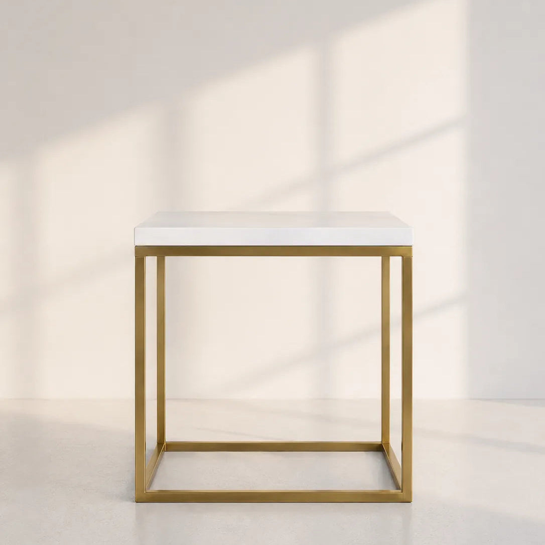 Image of A modern cube-shaped side table with a white marble top and a gold-finished metal frame, set against a minimalist off-white wall with soft window shadow patterns.