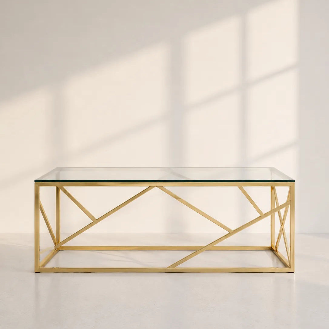 Image of A modern rectangular coffee table with a clear glass top and a geometric gold metal frame, set against a minimalist off-white wall with soft window shadow patterns.