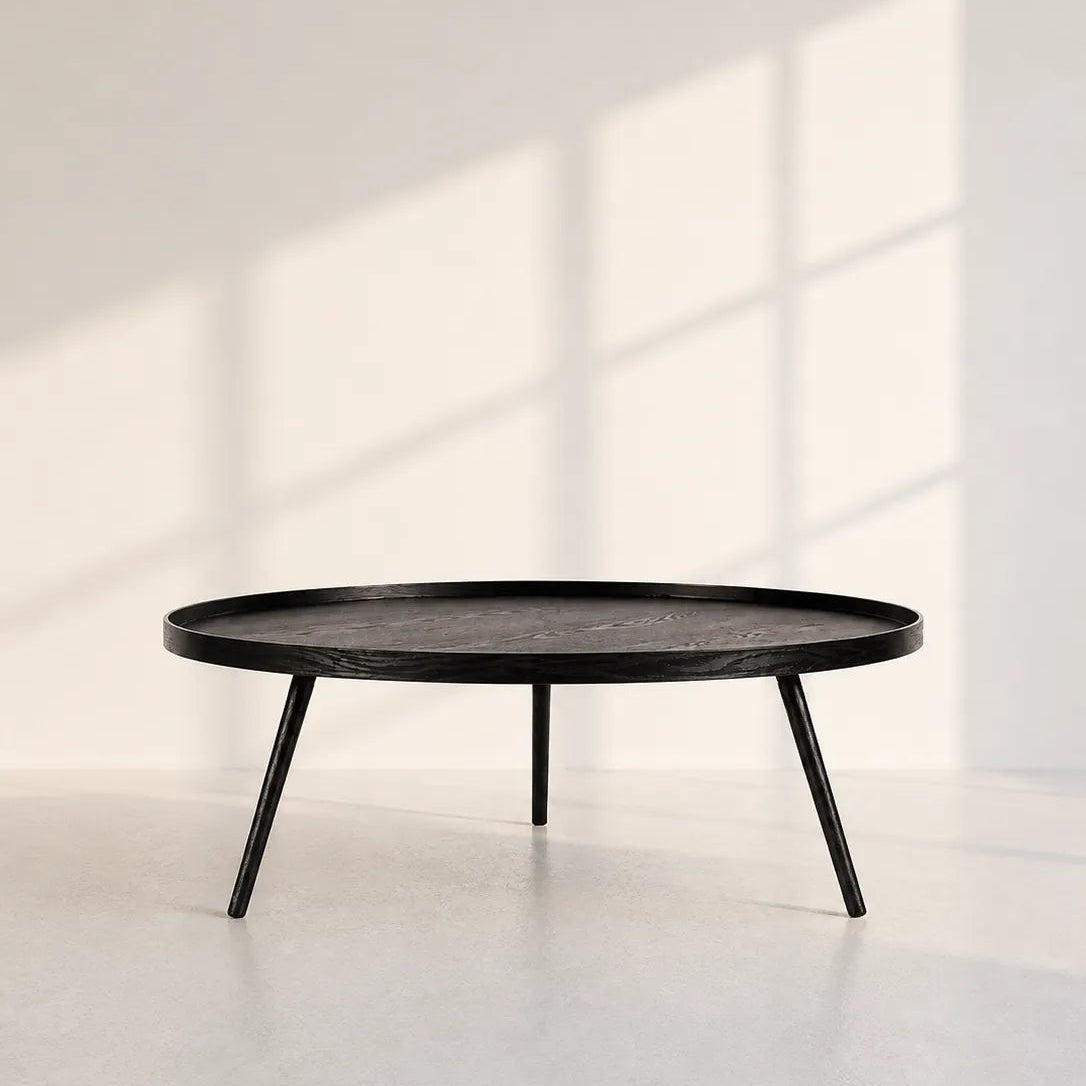Image of A round black coffee table with a dark wood-grain top and three tapered metal legs, set against a minimalist white wall with soft window shadow patterns.