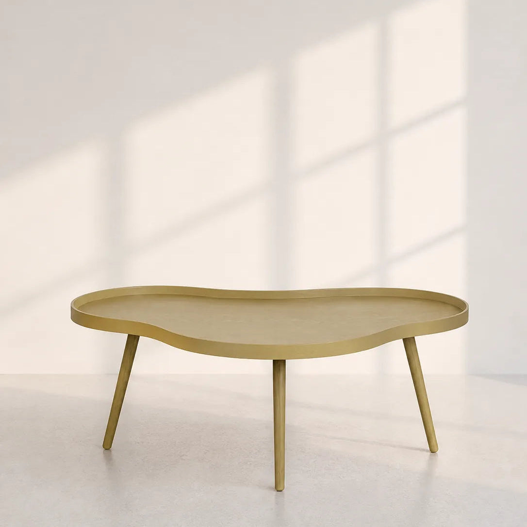 A modern organic-shaped coffee table with a light gold finish and tapered legs, set against a minimalist white wall with soft window shadow patterns.