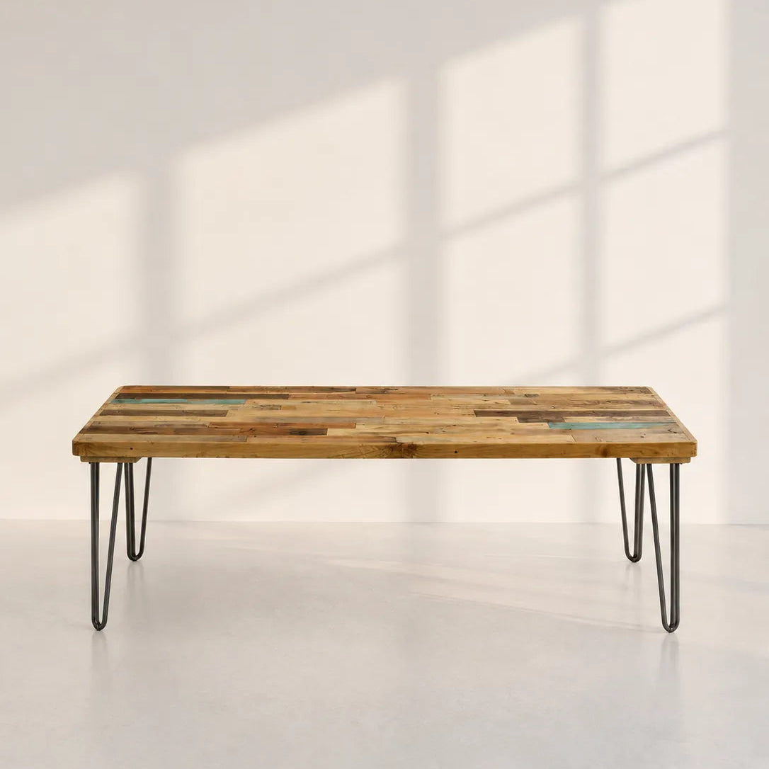 Image of A rectangular coffee table made from rustic, multi-toned reclaimed wood planks with black hairpin legs, set against a minimalist off-white wall with soft window shadow patterns.