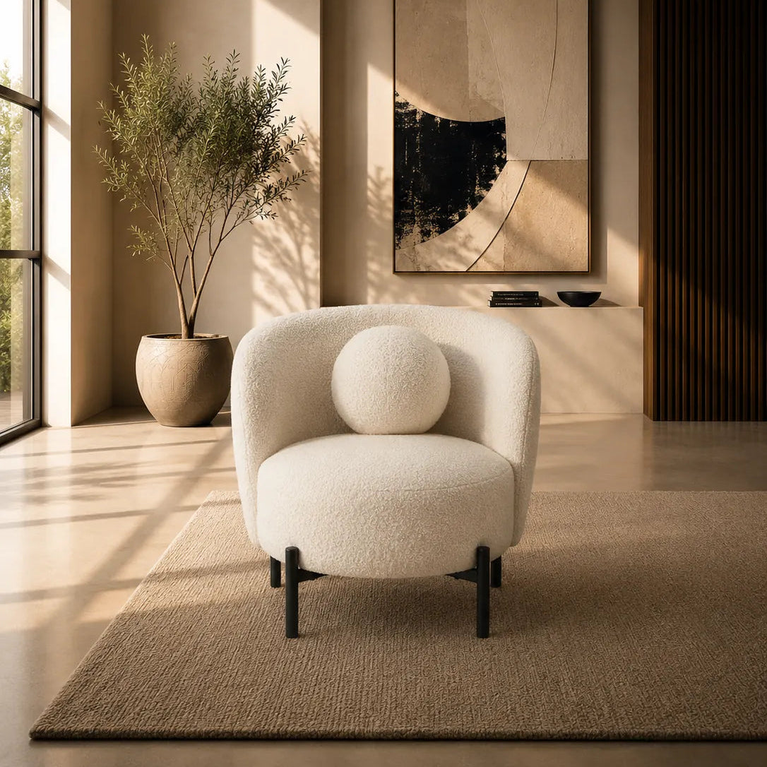 A modern cream boucle armchair with a round matching throw pillow sits on a textured beige rug in a warm, sunlit minimalist living room. The background features a large abstract art print, a potted plant, and warm ambient lighting.