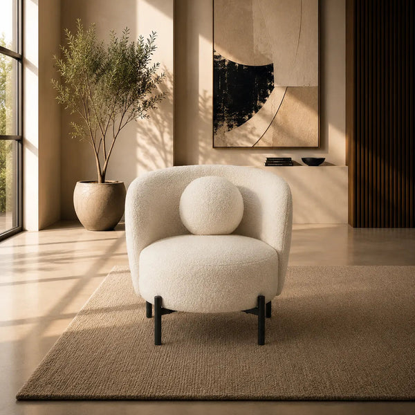 A modern cream boucle armchair with a round matching throw pillow sits on a textured beige rug in a warm, sunlit minimalist living room. The background features a large abstract art print, a potted plant, and warm ambient lighting.