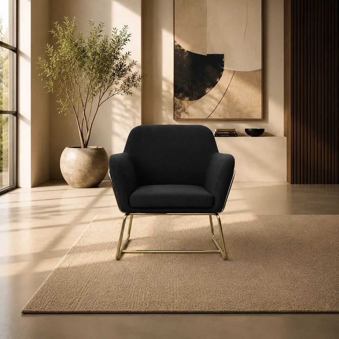 Image of A modern black upholstered accent chair with slim gold metal legs sits on a beige rug in a sunlit minimalist living room. A large abstract art print hangs on the wall behind it, with a potted plant to the left.