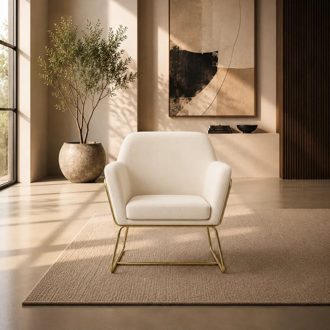 A modern white upholstered accent chair with a gold metal frame and legs, set against a warm beige wall with soft window shadow patterns.
