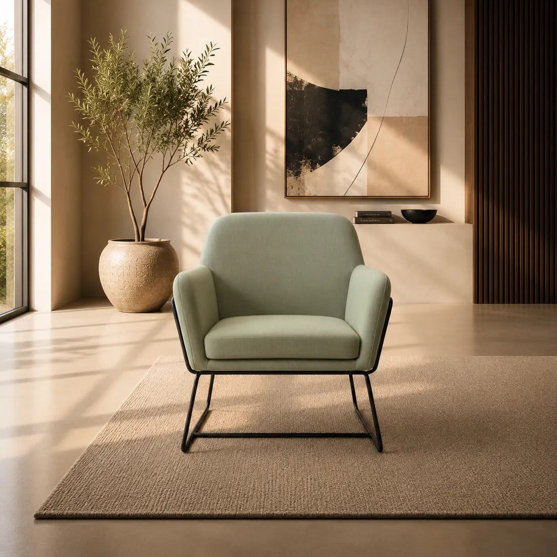 Image of A modern light sage green armchair with black metal legs sits on a textured beige rug in a minimalist, warm-toned living room. A large abstract art print hangs on the wall behind it, and a potted plant stands near the window on the left.